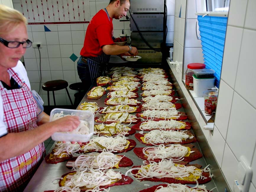 Auf einem Eisentisch liegen knapp 30 Fleischstücke die von einer Mitarbeiterin und dem Koch Christoph Hoffmann mit Gurkenscheiben Speck und Zwiebeln belegt werden.