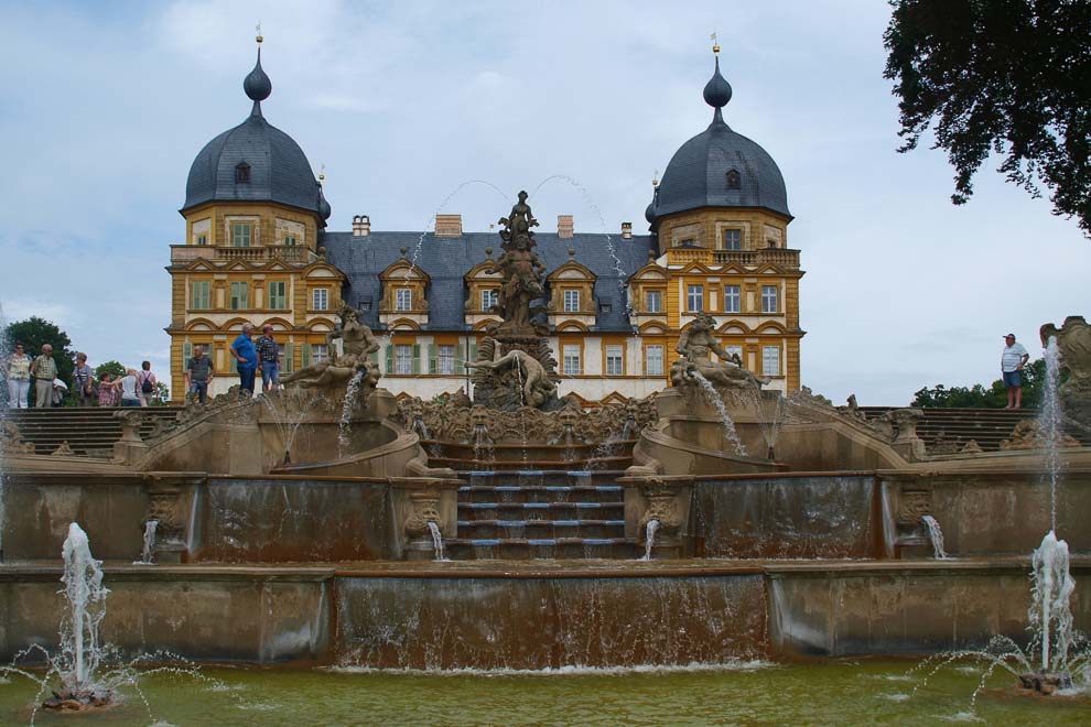 Das ockergelb-weiße Schloss steht hinter einem terrassenartig angelegten, mit vielen Skulpturen aus der Mythologie verzierten, sprudelnden Brunnen.